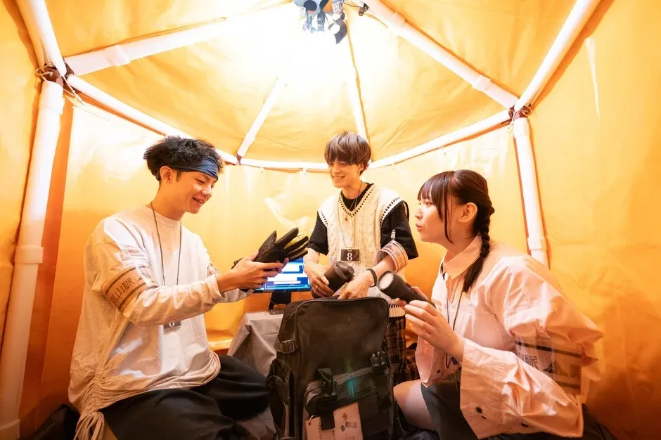 オレンジ色のテントのような空間に3人の若者が集まり、笑顔で会話をしています。男性はグローブを持ち、女性は懐中電灯を持っており、中央にはバックパックが置かれています。何らかのイベントやゲームに取り組んでいるような、楽しげな協調作業の様子がうかがえます。