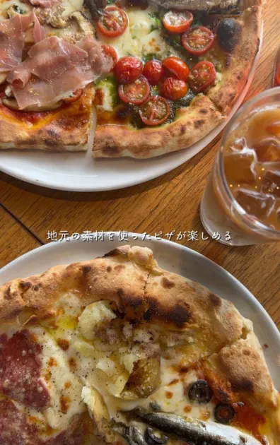 地元の素材を活かした2種類のピザが食欲をそそる写真です。生ハムとトマト、そしてサラミやアンチョビ、ポテトが乗ったピザが並び、美味しそうな食事の風景が広がっています。隣には飲み物も置かれています。