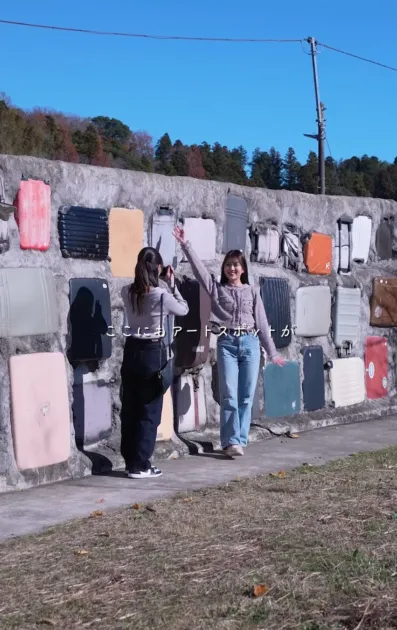 スーツケースが埋め込まれた壁面アートの前で、二人の女性が写真撮影を楽しんでいます。一人はポーズを取り、もう一人はカメラを構えています。晴れた日の屋外で、ユニークなアート作品を背景に思い出を作っている様子がうかがえます。
