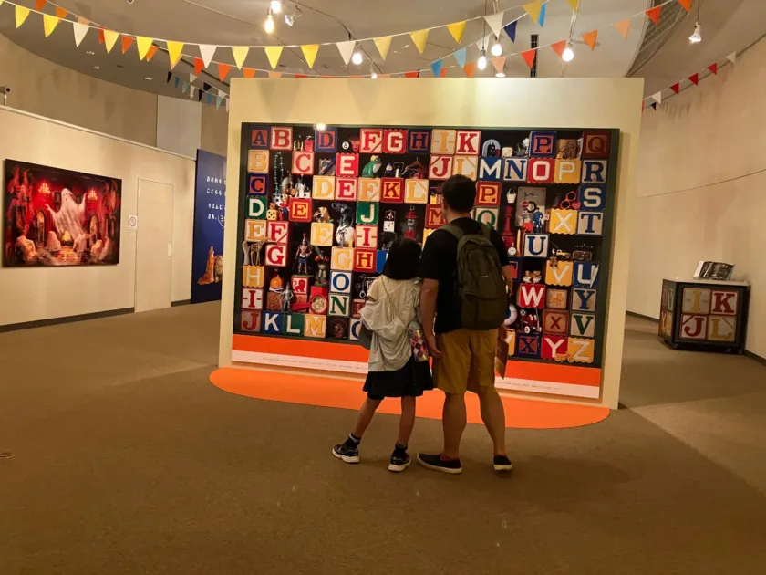 美術館で、父親と子供がアルファベットのブロックに様々な小物が飾られた大きな展示を鑑賞している様子