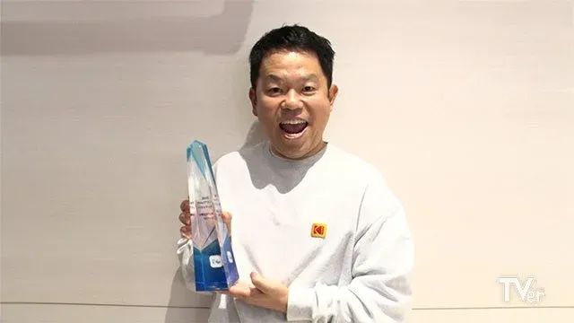 男性が「TVerアワード特別賞」のトロフィーを手に、満面の笑みを浮かべています。白いスウェットシャツにはKodakのロゴが見え、喜びを表現している様子です。