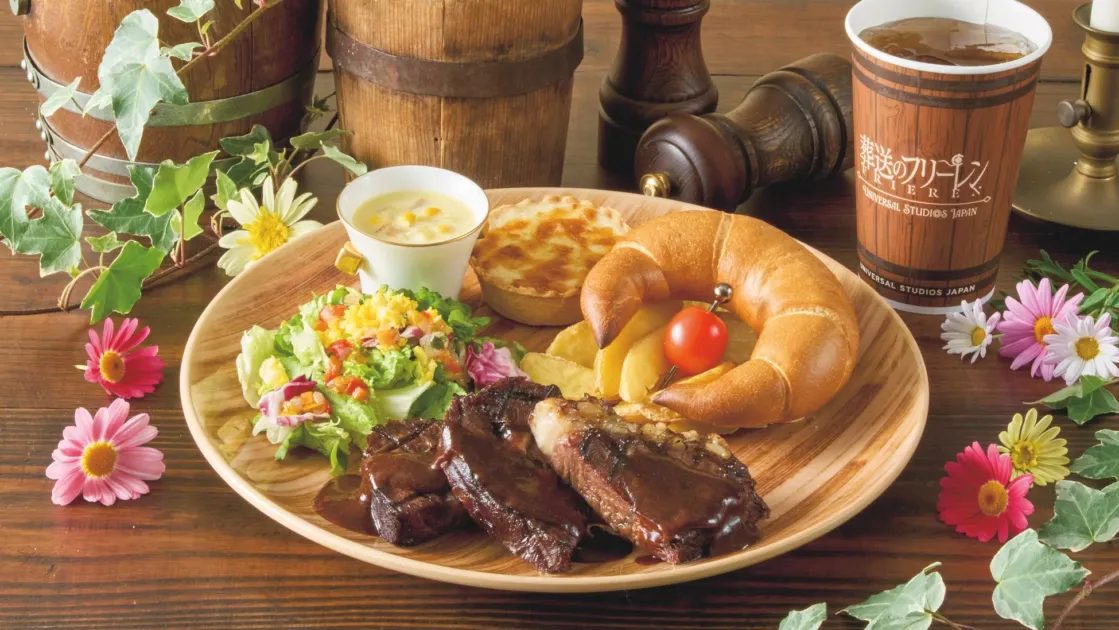 ユニバーサル・スタジオ・ジャパンの「葬送のフリーレン」コラボメニュー。肉料理、サラダ、クロワッサン、グラタン、ポテト、テーマドリンクが木製プレートに盛り付けられ、花や緑が添えられた豪華な食事セットです。