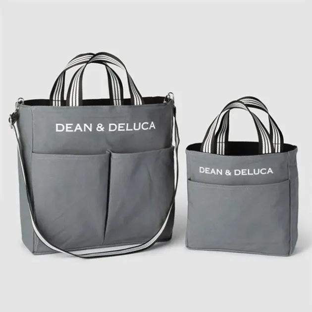グレーのキャンバス地トートバッグが大小2つ並べられています。どちらのバッグにも「DEAN & DELUCA」のロゴが白色で印字されており、持ち手は黒と白のストライプ柄です。