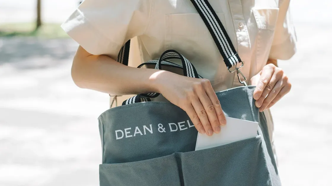 DEAN & DELUCAのグレーのトートバッグを持った人物が、バッグのポケットから白いものを出し入れしている様子を捉えたクローズアップ画像です。カジュアルな服装で、お出かけ中のワンシーンを思わせます。