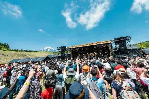 快晴の空の下、富士山を背景にした野外会場で、大勢の観客がステージ上のパフォーマンスを楽しむ音楽フェスの様子
