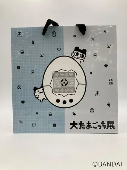 たまごっちのキャラクターとデバイスが描かれた、水色と白のツートンカラーの紙製ショッピングバッグです。レトロなピクセルアートのアイコンが散りばめられ、「大たまごっち展」の文字が見えます。