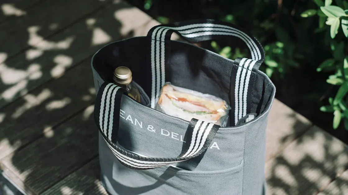 屋外の木製テーブルに置かれたDEAN & DELUCAのトートバッグ。バッグの中にはサンドイッチとボトルが入っており、ピクニックやランチの準備がされている様子がうかがえます。