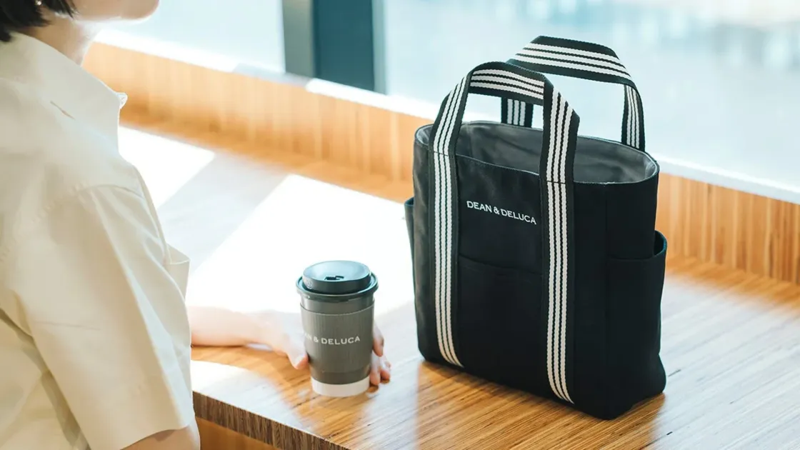 DEAN & DELUCAの黒いトートバッグとコーヒーカップが木製のテーブルに置かれ、隣に座る女性の手元が写っています。カフェでリラックスした時間を過ごしているような、スタイリッシュな日常のシーンです。