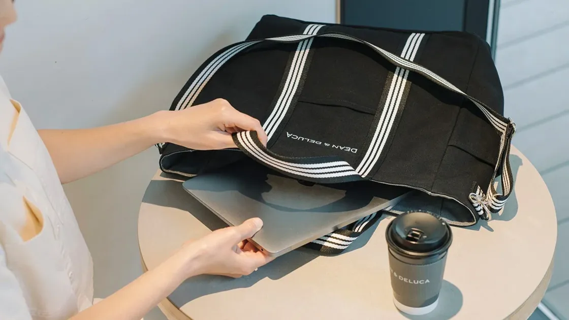 女性が黒いDEAN & DELUCAのトートバッグにノートパソコンを出し入れしている様子。隣には同ブランドのコーヒーカップが置かれており、仕事中の休憩や移動中のシーンを思わせる一枚です。