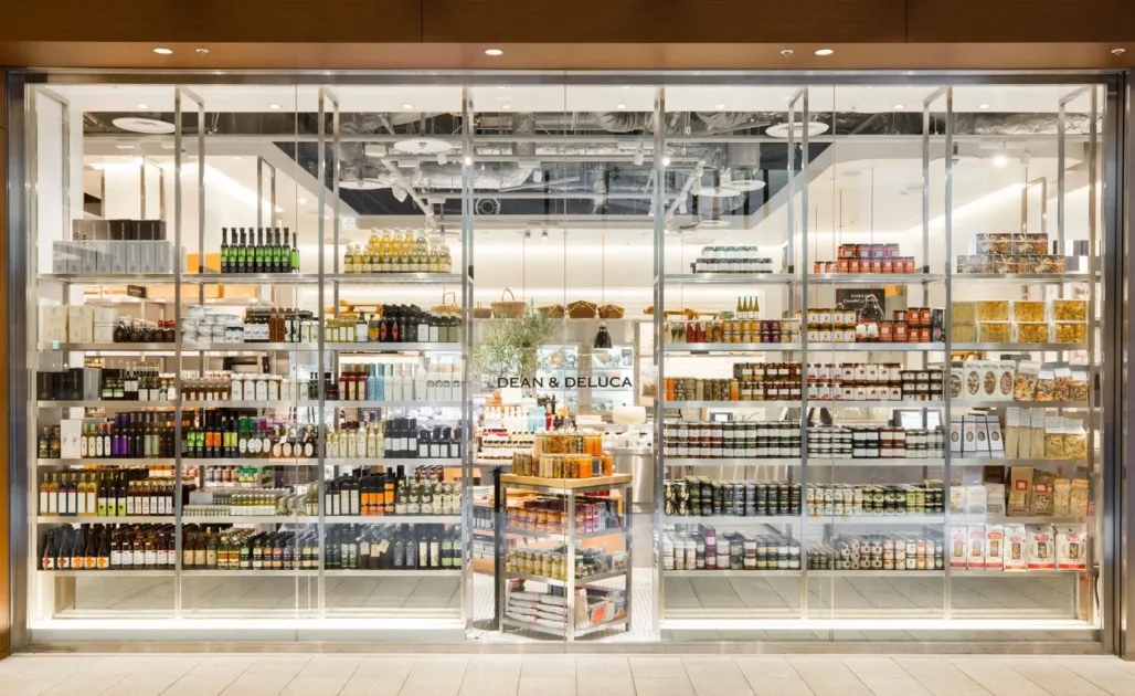 DEAN & DELUCAの店舗正面の様子。ガラス張りのショーウィンドウ越しに、色とりどりの瓶詰やパッケージ商品など、多種多様なグルメ食品が整然と並べられた商品棚が広がる。高級感のある食料品店の雰囲気が伝わる。