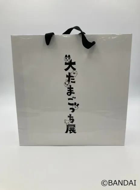 白地のシンプルな紙袋で、中央に「大たまごっち展」と書かれたロゴが縦に配置されています。文字の一部はたまごっちのキャラクターで表現されており、可愛らしい印象を与えます。