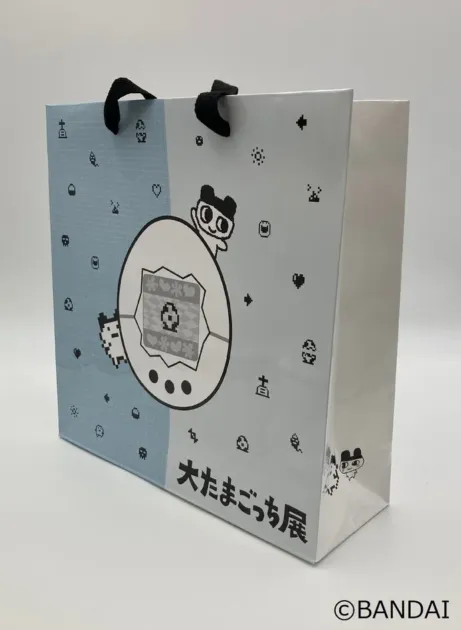 「大たまごっち展」のロゴが入った紙袋です。レトロなピクセルアートで描かれたたまごっちのキャラクターやゲーム画面がデザインされており、水色と白の2トーンカラーが特徴です。黒い持ち手が付いています。