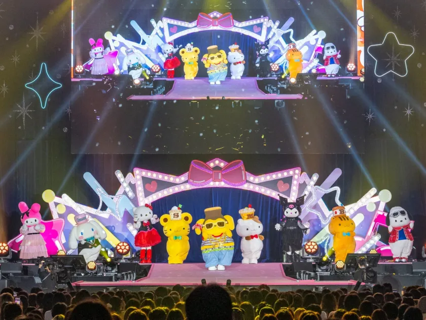 サンリオの人気キャラクターたちが華やかなステージでパフォーマンスを披露しているライブイベントの様子。カラフルな照明と装飾が特徴で、多くの着ぐるみキャラクターが並び、観客がそのショーを楽しんでいる。
