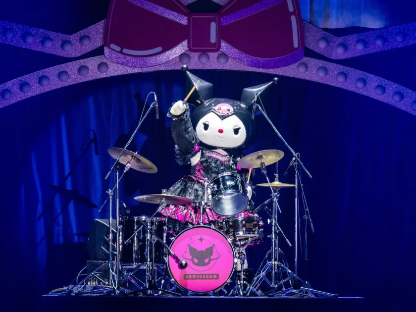サンリオの人気キャラクター、クロミがステージでドラムを演奏している様子です。黒とピンクの衣装をまとい、背景には大きなリボンの装飾があります。バスドラムには「#世界クロミ化計画」と書かれています。