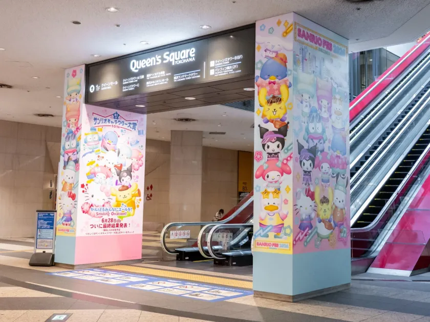 横浜のクイーンズスクエア入口付近で撮影された、サンリオキャラクター大賞2024とSANRIO FES 2024のカラフルな広告が施された柱が目立つ写真です。案内表示やエスカレーターも見え、多くのサンリオキャラクターが描かれた賑やかなイベント告知が目を引きます。