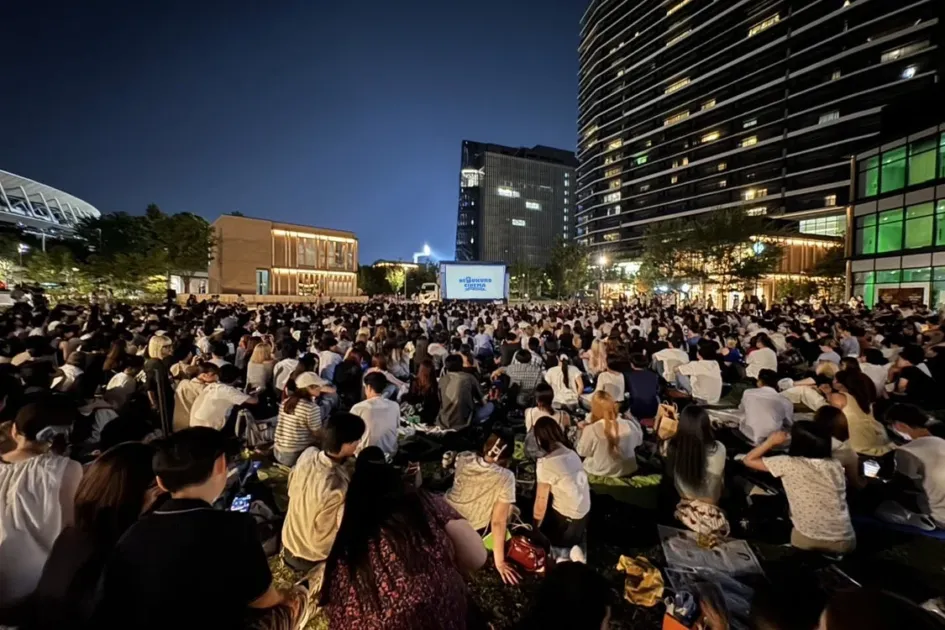夜の都市公園で、多くの人々が芝生に座り野外映画を楽しむ様子