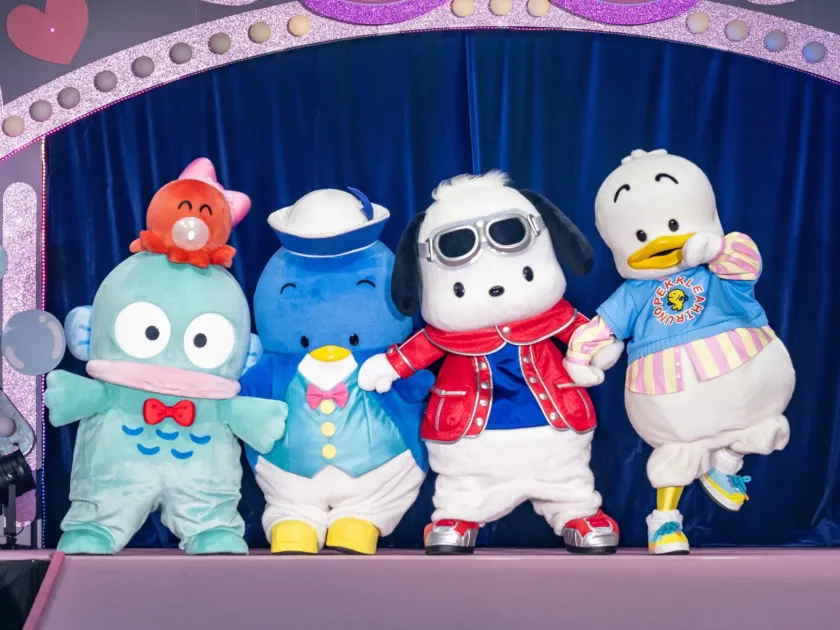 青いカーテンを背景にしたステージに、サンリオの人気キャラクターであるハンギョドン、タキシードサム、ポチャッコ、アヒルのペックルの着ぐるみが並んでポーズをとっています。イベントやショーの一場面を捉えた、可愛らしく華やかな写真です。