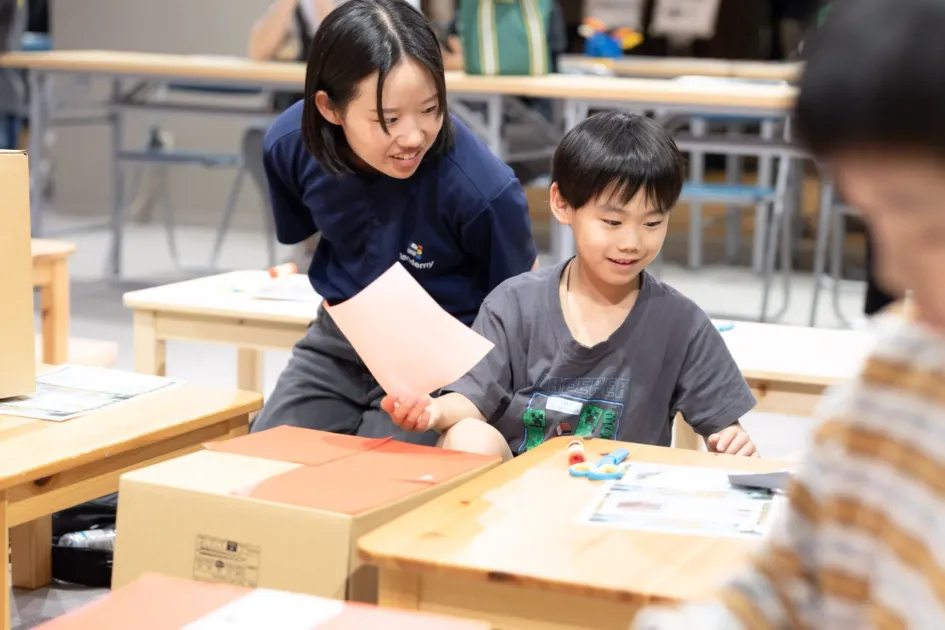 室内で、年上の女の子と年下の男の子がテーブルを囲み、何か作業をしている様子です。女の子はピンクの紙を持ち、男の子は笑顔で手元の物を見ています。学習やワークショップのような雰囲気で、楽しそうに交流しています。