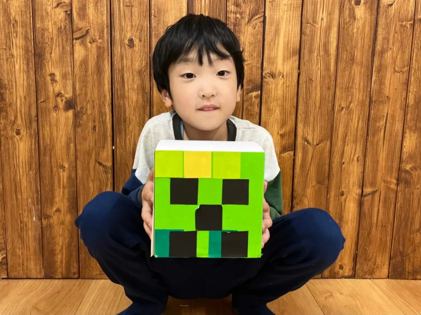 木目の壁を背景に、マインクラフトのクリーパーの顔を模した緑色の箱を抱えてしゃがむ男の子の姿です。カメラに向かって微笑んでいます。