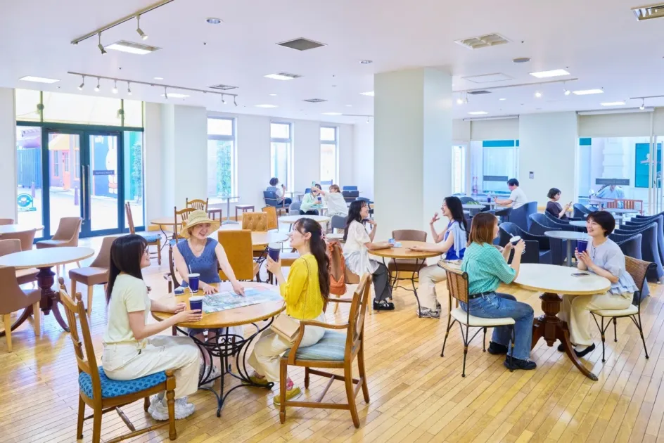 明るく広々としたカフェまたはラウンジで、多くの人々が談笑したり、地図を見たりしながらくつろいでいる様子