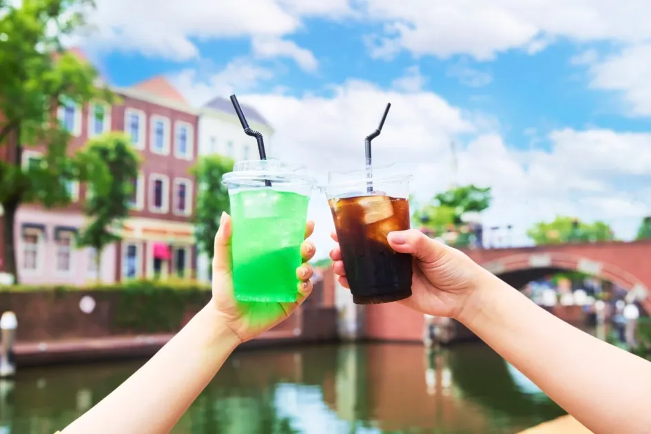 晴れた日に、水辺の美しい街並みを背景に、メロンソーダとアイスコーヒーで乾杯している様子