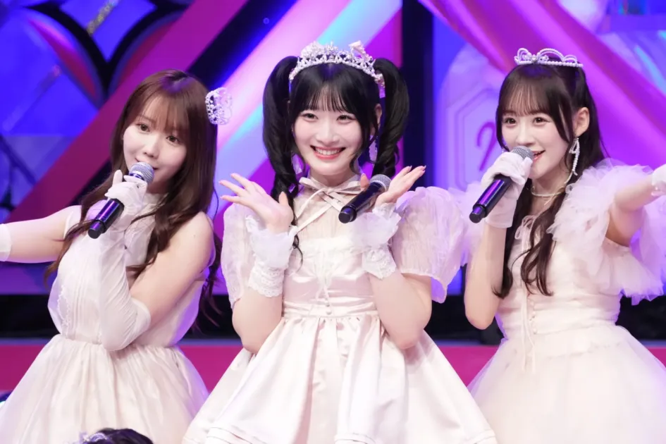 明るい照明のステージで、可愛らしいドレスを着た3人の女性アイドルがマイクを持って歌唱している。