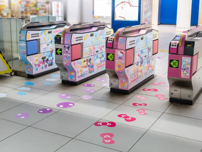 駅の改札口がサンリオキャラクターでカラフルに装飾されています。ハローキティやクロミなどが描かれた自動改札機が並び、床にはキャラクターをモチーフにした足跡や模様がデザインされており、可愛らしい雰囲気を作り出しています。