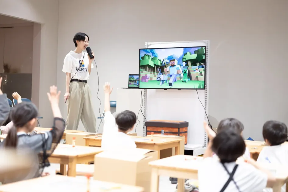講師がマイクを使い、マインクラフトの画面を映しながら子供たちに話しかける教育イベントの様子。子供たちは積極的に手を挙げており、活発な学びの場であることが伝わります。