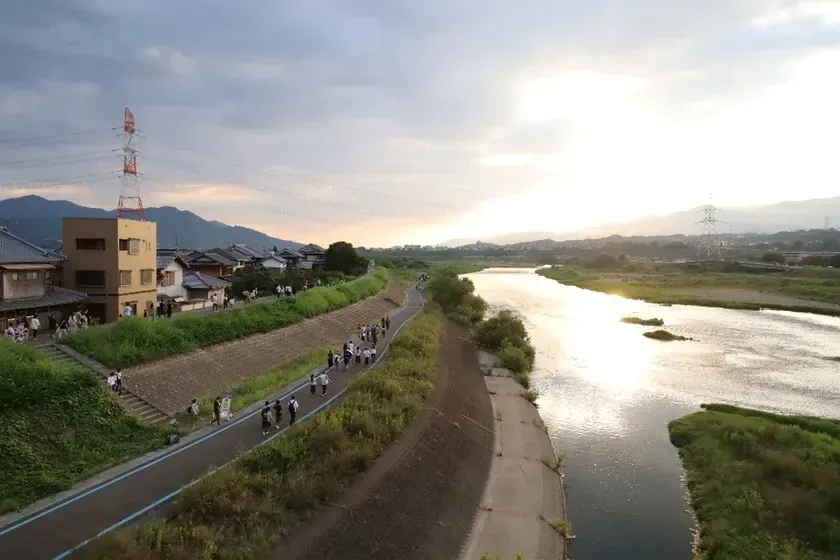 夕暮れ時の川沿いの風景。多くの人々が河川敷の道を散歩しています