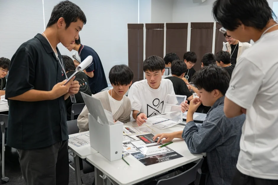 複数の若い男性が教室のような場所で、テーブルを囲んでグループワークやディスカッションを行っている様子