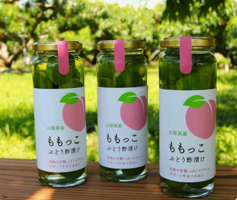 山梨県産の若桃をぶどう酢で漬けた「ももっこ」の瓶が3本、緑豊かな屋外に並べられています。