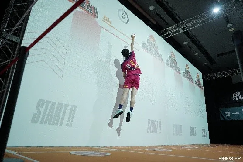 スポーツウェアを着た人物が、巨大スクリーンに映し出された「最高到達点!!」の表示に向かって高くジャンプしている