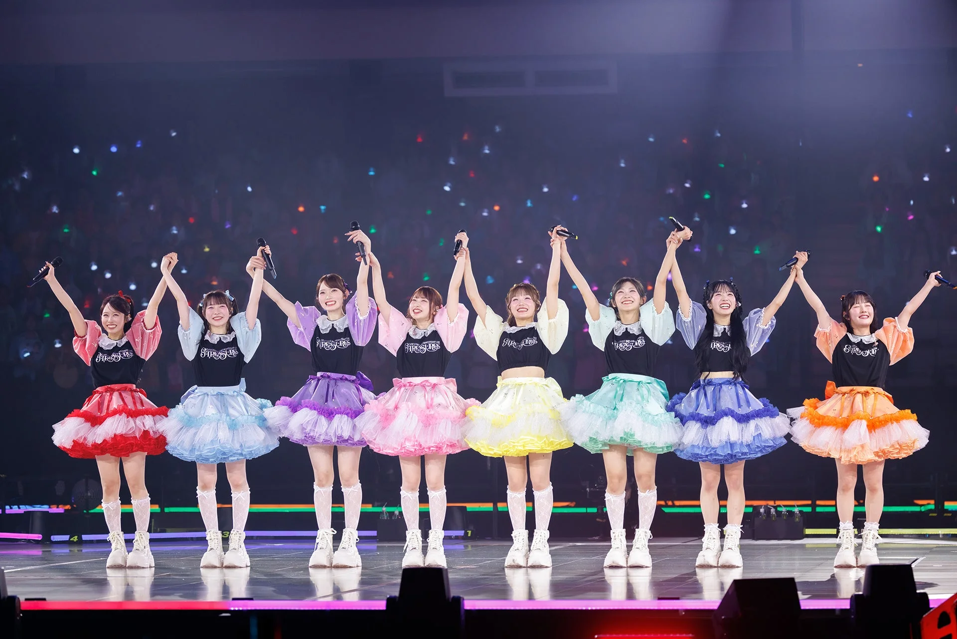 8人組の女性アイドルグループがステージ上で手を繋ぎ、腕を上げて笑顔で観客に手を振っている様子。