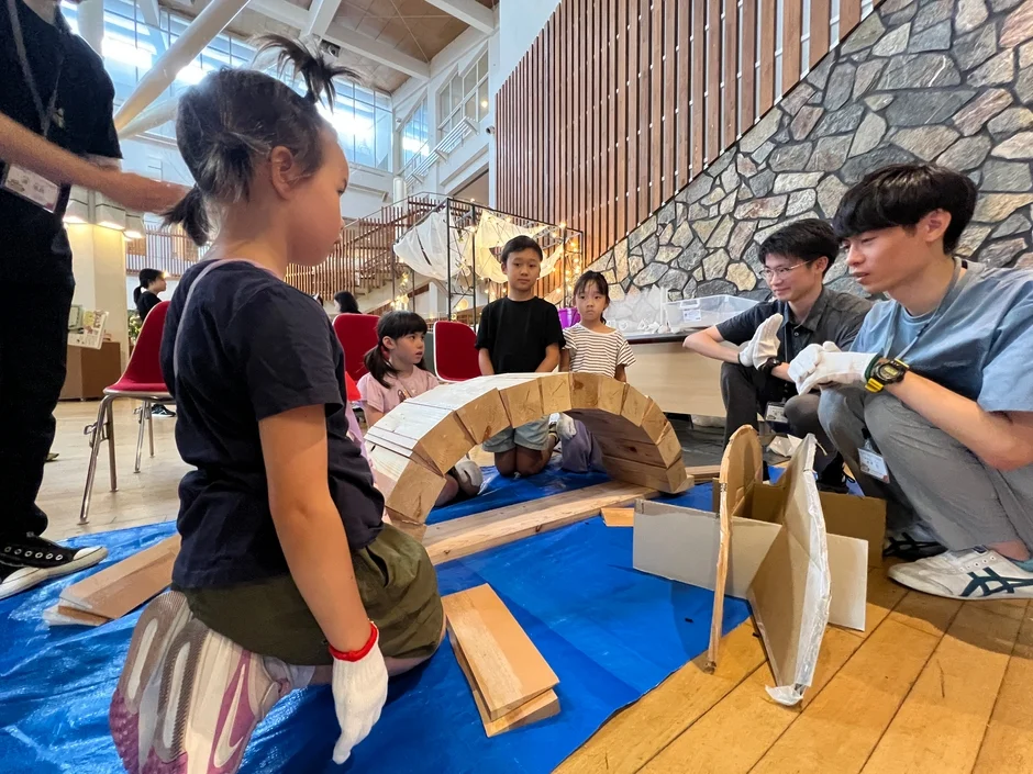 屋内施設で子供たちが木材を使った建築ワークショップに参加しています。大人が見守る中、子供たちは協力して木製のアーチ構造物などを制作しており、創造的な体験を楽しんでいる様子が伝わります。