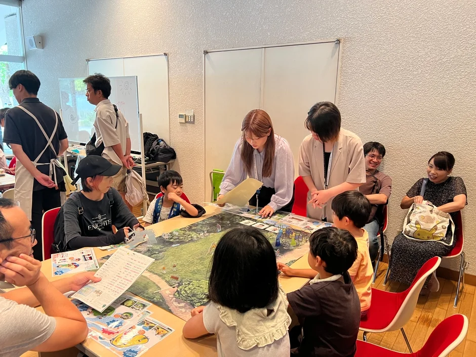 室内で大人と子供たちがテーブルを囲み、ボードゲームや教育的な活動を楽しんでいる様子。参加者は笑顔で交流しており、熱心に活動に参加している。ポケモン関連のアイテムも見える。