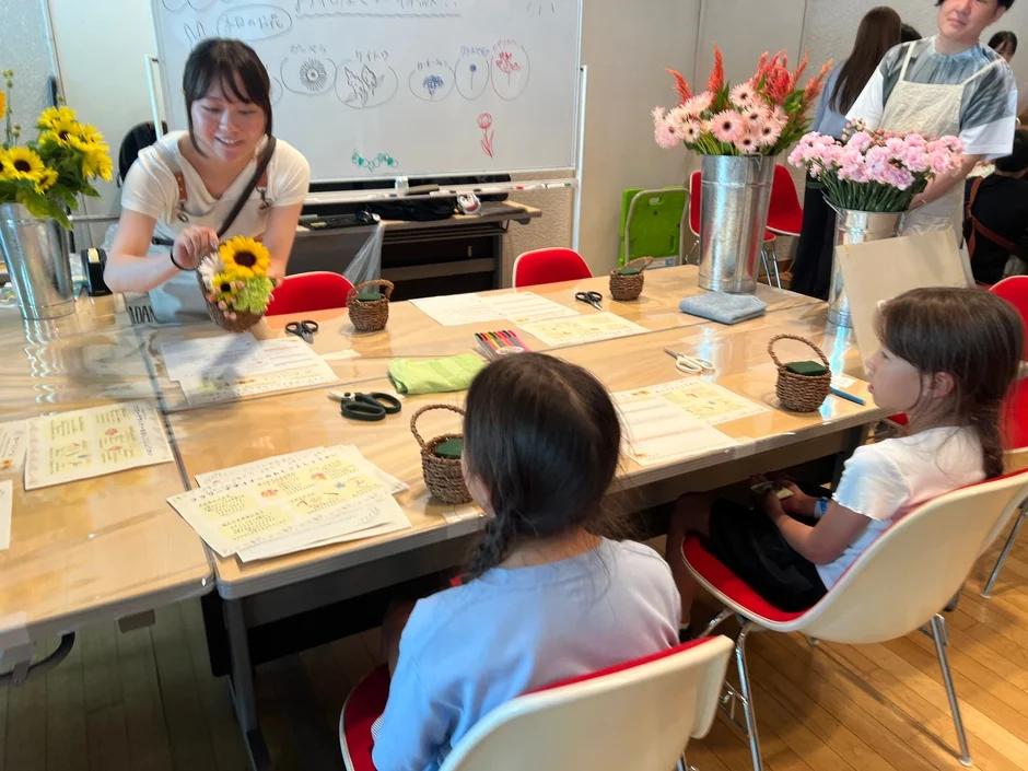 女性が講師となって子供たちにフラワーアレンジメントを教えているワークショップの様子です。テーブルには様々な花や道具が並び、明るく楽しい雰囲気です。