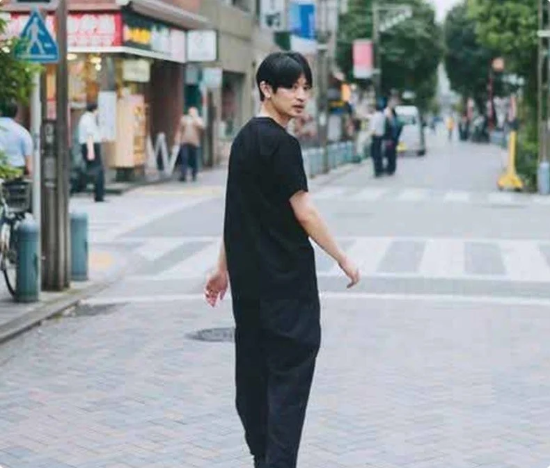 黒いTシャツとパンツを着用した若い男性が、街中の舗道を歩きながら後ろを振り返っている様子を捉えた写真です。