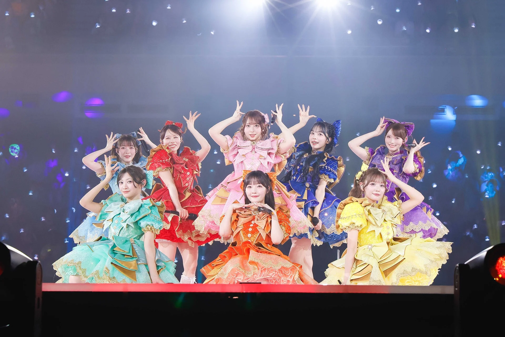 華やかなステージで、カラフルなフリル衣装を身につけた9人の女性アイドルグループが笑顔でポーズをとっています。