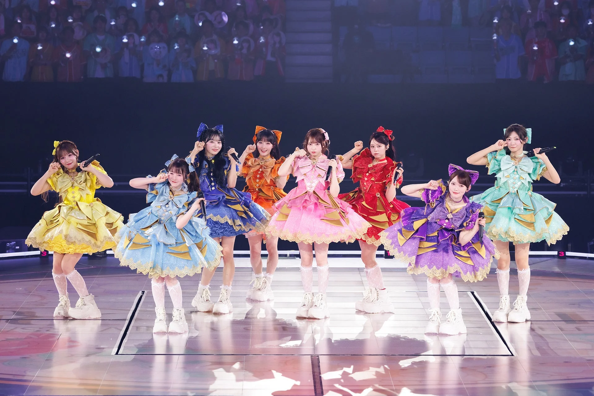 華やかなフリルのドレスをまとった8人の女性アイドルがステージでパフォーマンスしている様子。背景にはペンライトを持つ大勢の観客が見える。