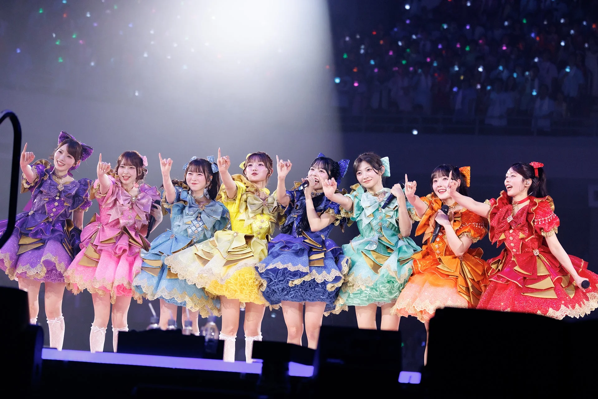 カラフルなアイドル衣装をまとった8人の女性グループが、ステージで笑顔でパフォーマンスしているライブの様子。
