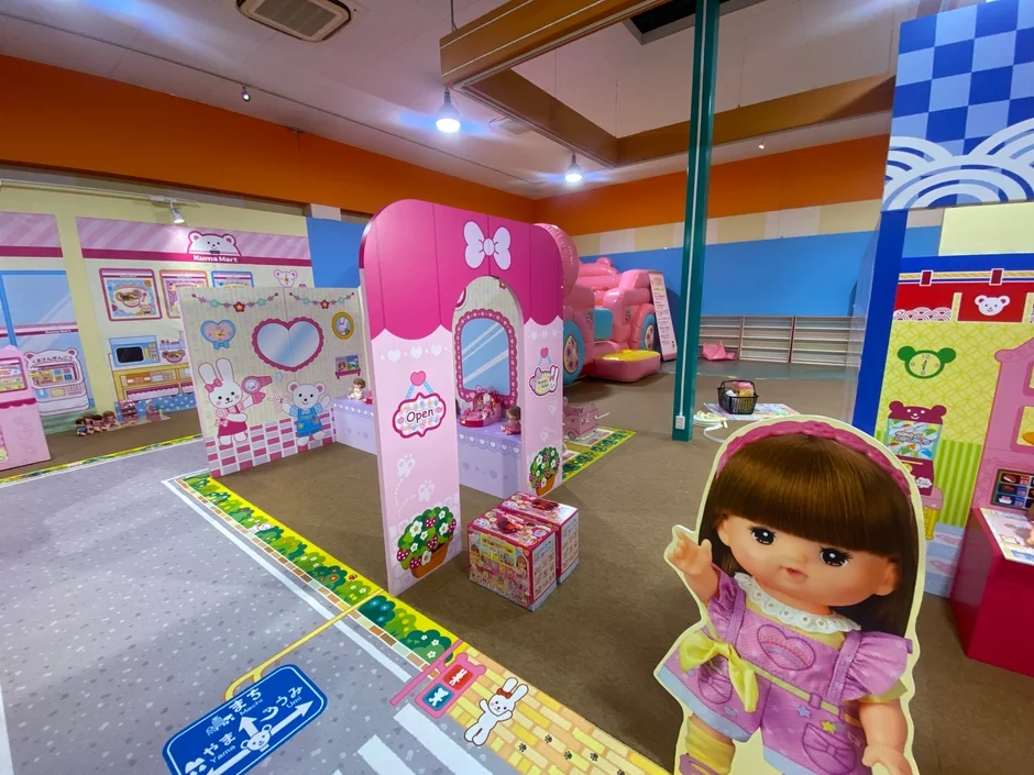 カラフルな室内プレイルームで、子供向けの遊び場が広がっています。「Kuma Mart」という名前のスーパーマーケットやピンクのドレッサーエリアがあり、大きな人形や動物のキャラクターのパネルが配置されています。