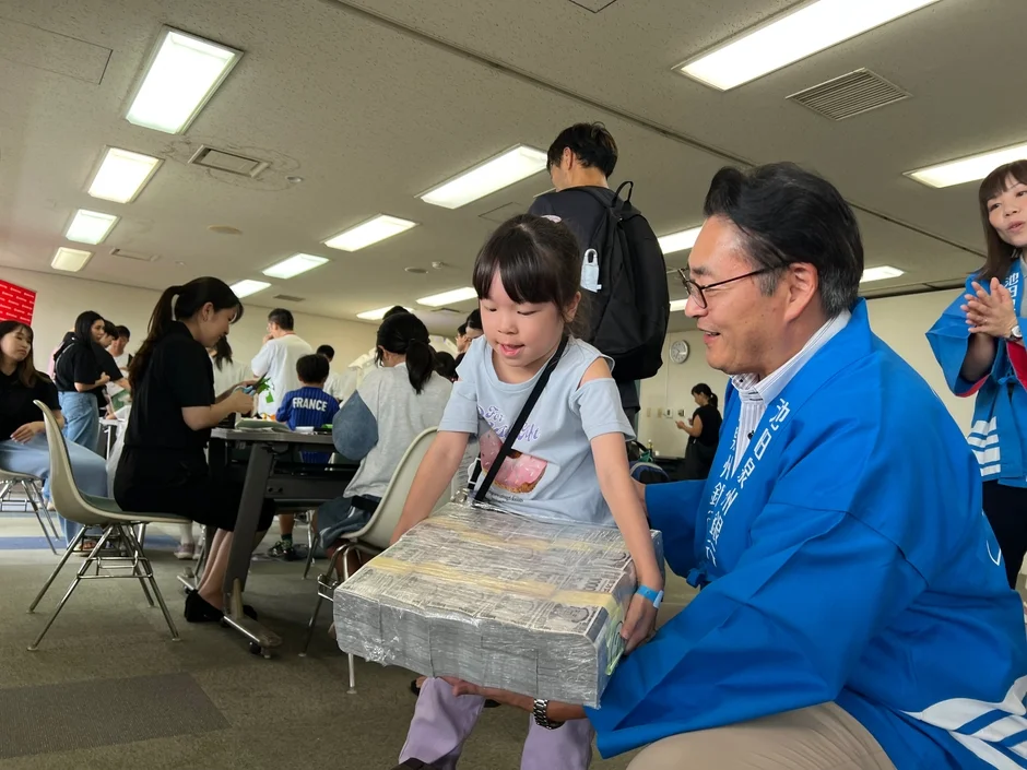 青い法被を着た男性が、大量の紙幣の束のようなものを運ぶ女の子を笑顔で手伝っている様子です。屋内のイベント会場で、多くの人々が活動に参加している賑やかな一場面で、地域のお祭りや体験イベントのようです。