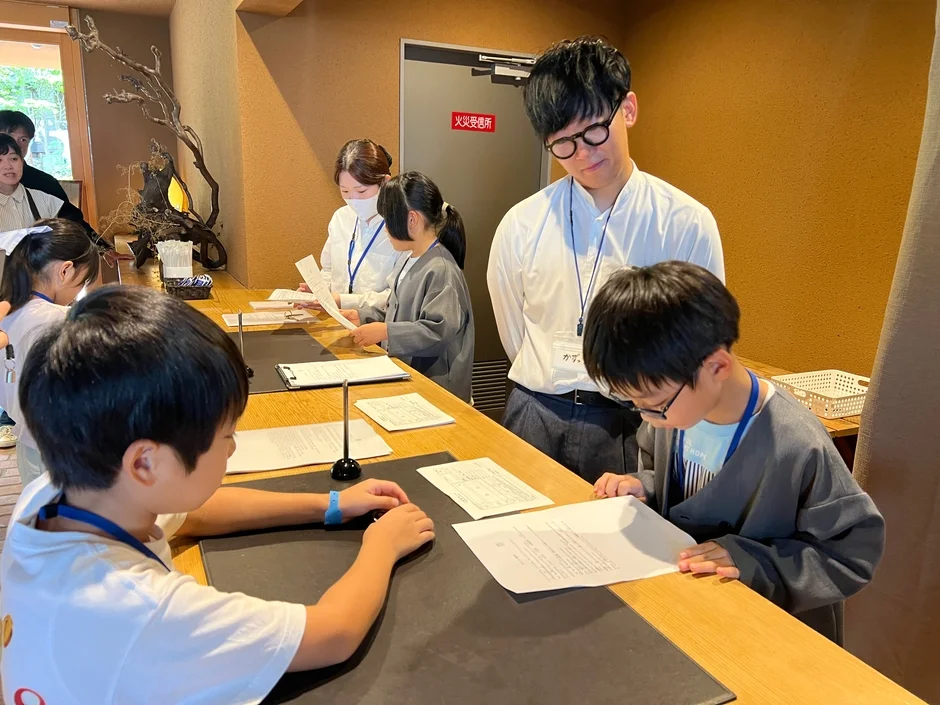 子供たちが大人と一緒にカウンターで書類に目を通したり、手続きを行ったりしている様子。イベントの受付や体験学習の開始前のような雰囲気で、数人の子供と大人が写っています。