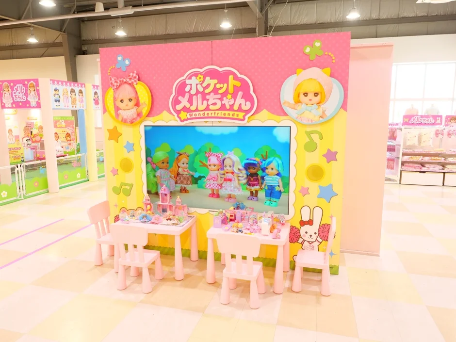 「ポケットメルちゃん Wonderfriends」のカラフルなディスプレイが中央にあり、ピンクのテーブルと椅子が置かれた子供向けの遊び場またはイベントブースの様子です。