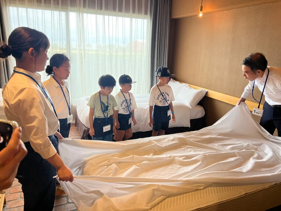 ホテルの一室で、子供たちがスタッフからベッドメイキングの指導を受けている。大人たちがシーツを広げる作業を実演し、子供たちは真剣な表情でその様子を見つめている。職業体験プログラムの一場面。