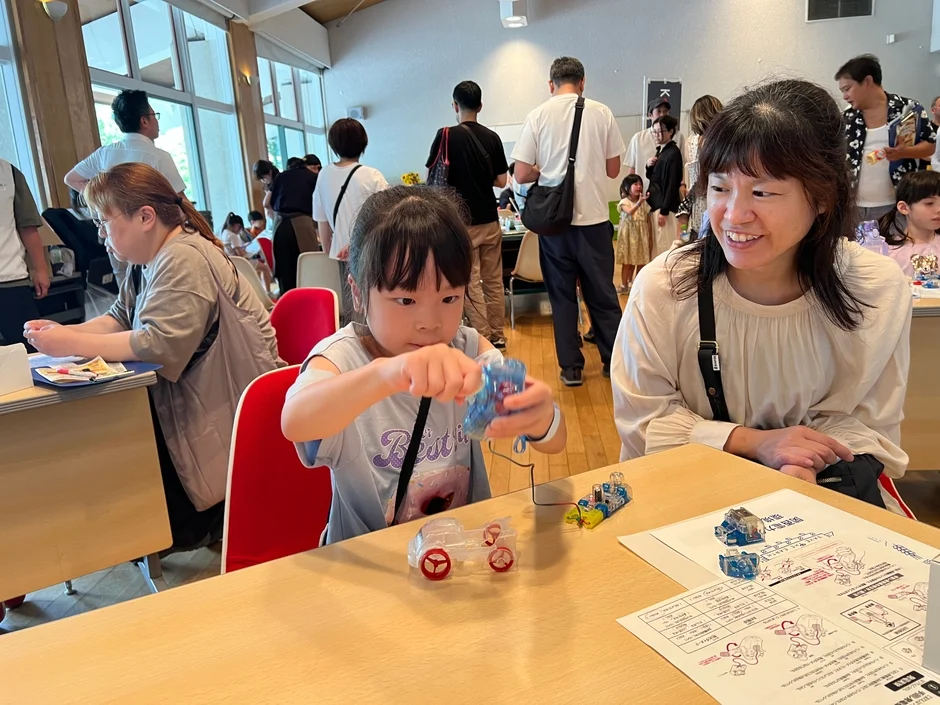 明るい室内で、親子が工作ワークショップに参加している様子。幼い女の子が透明なプラスチック製のおもちゃを組み立てており、隣に座る女性が笑顔で見守っている。テーブルには組み立て説明書が広げられている。