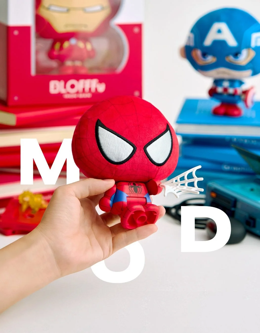 手がスパイダーマンのぬいぐるみを持ち、背景にはアイアンマンとキャプテンアメリカのフィギュアが並べられています。マーベルキャラクターグッズのコレクションをテーマにした写真で、大きな白い文字「MSD」がオーバーレイされています。