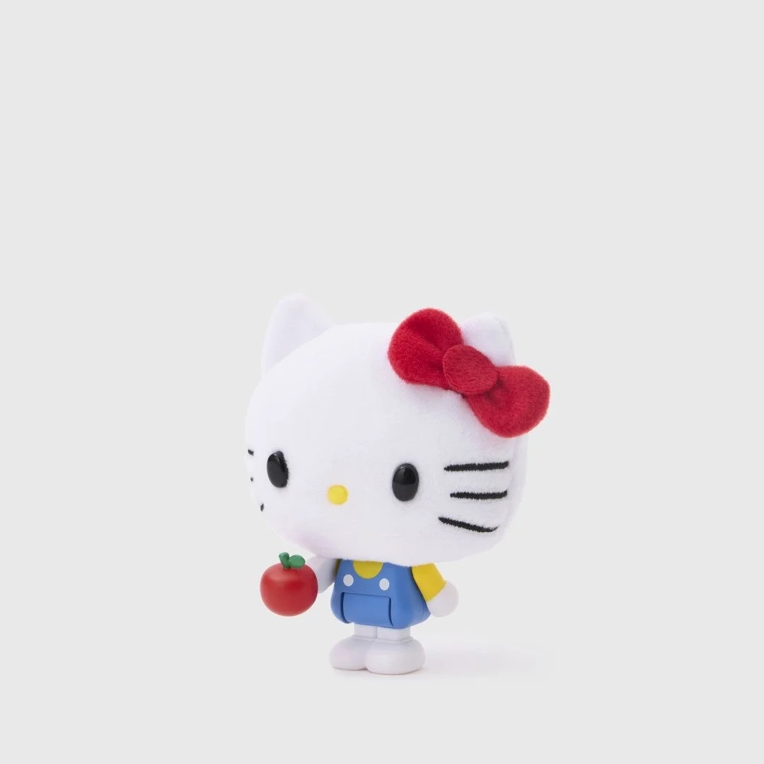白い背景に立つハローキティのフィギュアです。赤いリボンをつけ、青いオーバーオールを着用し、左手には小さな赤いリンゴを持っています。可愛らしい姿が特徴です。