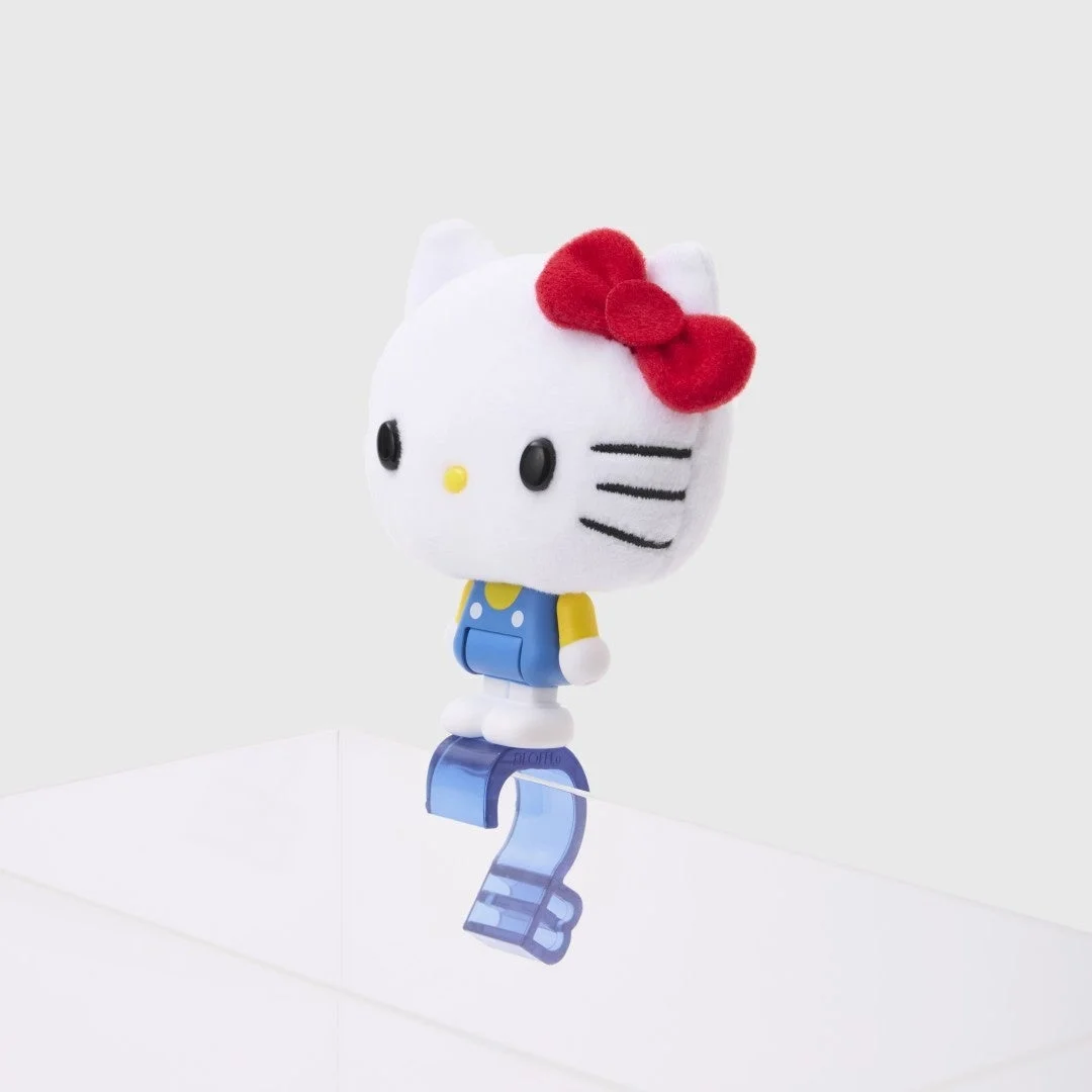 白い背景に、青いオーバーオールを着て赤いリボンをつけたハローキティのフィギュアが、透明な青いクリップにぶら下がっている様子。